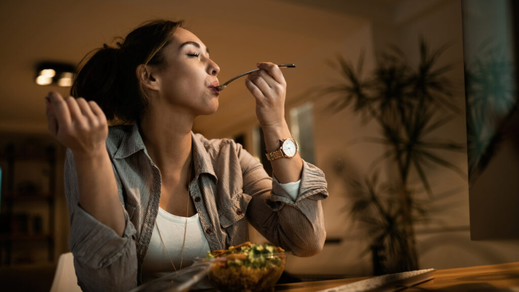 low-angle-view-young-woman-eating-salad-with-her-eyes-closed-while-using-computer-night-home
